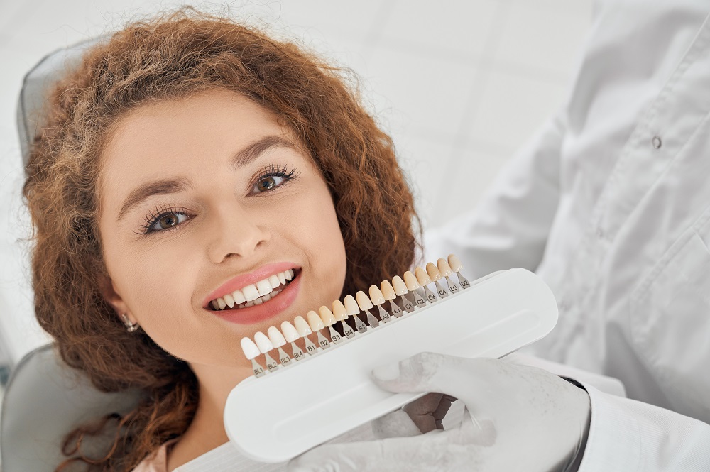 Closeup of beautiful young woman lying on dental chair, looking at camera and smiling while male dentist keeping in hands teeth color range. Girl doing whitening procedure in dental office.